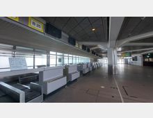 Leere Check-in-Schalter in einem Flughafenbereich mit grauem Boden und Decke. Im Hintergrund sind große Fensterfronten und Beleuchtungsschilder mit Bereichsangaben wie "B 25" sichtbar.