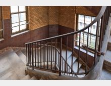 Treppenhaus mit gebogener Metallgeländer in einem Gebäude mit gelblich-braunen Ziegelwänden und großen Fenstern. Die Fliesen an den Wänden unterhalb der Fenster sowie die breite Fensterfront lassen Tageslicht in den Raum.