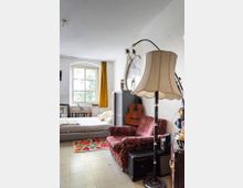 Wohnzimmer mit einer gemütlichen Einrichtung: Im Vordergrund steht ein roter Sessel mit Blumenmuster neben einer Stehlampe mit großem Stoffschirm. Im Hintergrund befinden sich ein Bett, ein gelber Vorhang vor einem Fenster, ein Bücherregal, eine Akustikgitarre und ein kleiner Schrank.