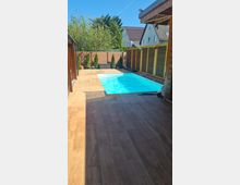 Privater Außenbereich mit rechteckigem Swimmingpool, umgeben von Holzterrassenboden und Sichtschutzwänden aus Holz. Im Hintergrund sind Bäume, Sträucher und ein Wohnhaus erkennbar.