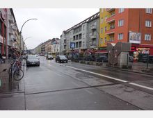 Straßenszene in einer Stadt mit mehrstöckigen Wohn- und Geschäftshäusern, darunter graue, gelbe und orangefarbene Fassaden. Im Vordergrund stehen Fahrräder auf einem Gehweg, Autos fahren auf der nassen Fahrbahn, und ein U-Bahn-Schild weist auf eine Haltestelle hin.