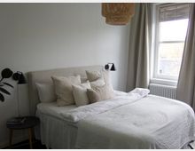 Schlafzimmer mit einem Doppelbett mit weißer Bettwäsche und mehreren beigen Kissen. Links neben dem Bett steht ein kleiner runder Beistelltisch mit einer Lampe, und rechts befindet sich ein Fenster mit hellen Vorhängen sowie einem Heizkörper darunter.