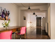 Ein heller Innenraum mit Holzboden, weiß gestrichenen Wänden und Hängeleuchten an der Decke. Im Vordergrund steht ein Holztisch mit pinkfarbenen Stühlen, darauf eine Vase mit Tulpen; weiter hinten befinden sich Wände mit Kunstbildern, Türen und ein Kleiderständer.