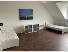 Schlafzimmer mit dunklem Holzfußboden, zwei weißen Einzelbetten auf gegenüberliegenden Seiten, einem weißen Regal in der Mitte und zwei kleinen, goldenen Beistelltischen. An der weißen Wand hängt ein großes Bild eines Fernsehturms bei Dämmerung.