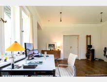 Ein heller Raum mit einem großen Schreibtisch vor bodentiefen Fenstern auf der linken Seite. Auf dem Schreibtisch stehen ein Computerbildschirm, eine gelbe Tischlampe und Schreibutensilien. Im Hintergrund befinden sich eine weiße Tür, ein Sideboard mit Kunstobjekten und eine Fitnessmaschine an der rechten Wand.