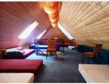 Ein Dachraum mit holzvertäfelten Wänden und zwei länglichen Dachfenstern, die Licht hereinlassen. Der Raum ist mit Matratzen, Sitzkissen und Stühlen eingerichtet, wobei eine Wendeltreppe mit Geländer in der Mitte sichtbar ist.