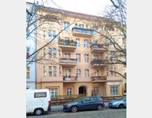 Mehrstöckiges Wohnhaus mit hellbeiger Fassade, roten Ziegeldach und mehreren Balkonen mit schmiedeeisernen Geländern. Im Vordergrund stehen geparkte Autos an einer Kopfsteinpflasterstraße, teilweise verdeckt von kahlen Baumästen.