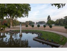 Parklandschaft mit einem kleinen Teich im Vordergrund, der von Blumenbeeten und einem geschwungenen Weg umgeben ist. Im Hintergrund sind eine große Wiese mit Baumreihen, ein einzelner Baum in der Mitte und ein angedeutetes Gebäude mit Kuppel in leichter Nebelstimmung zu sehen.