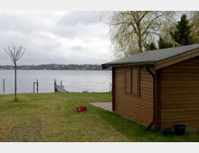 Holzhaus mit Flachdach am Ufer eines Sees, daneben eine Rasenfläche mit einem jungen Baum und einem kleinen Steg, der ins Wasser führt. Im Hintergrund sind bewaldete Ufer und Gebäude auf der gegenüberliegenden Seeseite sichtbar.
