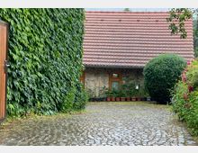 Gebäuderückseite mit Ziegeldach und Steinmauer, umgeben von Efeu, einem großen Strauch und Blumen. Ein gepflasterter Hof führt zum Eingang, der von Blumen in Töpfen dekoriert ist.