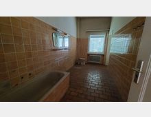 Ein Badezimmer mit orange-braunen Fliesen an Wänden und Boden, einer eingebauten Badewanne links, zwei Spiegeln über einem Waschbeckenbereich und einem Fenster mit Jalousien im Hintergrund. Ein Heizkörper und eine Toilette befinden sich unter dem Fenster auf der rechten Seite.