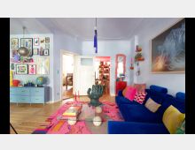 Ein Wohnzimmer mit hellblauen Wänden, einem blauen Sofa, bunten Kissen und einem pinken Teppich. Ein Glastisch mit einer skulpturalen Hand als Fuß steht im Raum, Wände sind mit vielen farbigen Bildern dekoriert, und im Hintergrund führt eine Türöffnung in einen weiteren bunt gestalteten Raum.