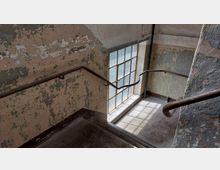 Verlassene Treppe in einem alten Gebäude mit abblätternder Wandfarbe und einem bodentiefen Metallfenster, durch das diffuses Tageslicht einfällt. Die Treppe hat dunkle Steintritte und geschwungene Metallgeländer.