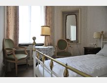 Kleines Schlafzimmer im klassischen Stil mit einem Einzelbett, einem Holzstuhl mit grünem Polster, einem Nachttisch mit Lampe und einem Tisch mit Spitzendecke und Stehlampe. Im Hintergrund ein Fenster mit gemusterten Vorhängen und ein Spiegel über einer Konsole.