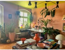 Wohnzimmer mit rosa Wänden und einer gelb gestrichenen Decke, eingerichtet mit zahlreichen Pflanzen in Töpfen und hängend an der Decke. Im Raum stehen ein Schreibtisch mit Stuhl vor einem Fenster, ein Couchtisch mit Büchern und Dekorationsartikeln sowie eine moderne Deckenleuchte.