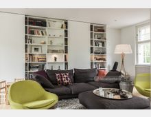 Wohnzimmer mit modernen Möbeln, darunter eine dunkelgraue Sofalandschaft, zwei hellgrüne Design-Sessel und ein runder Couchtisch mit Deko-Elementen. Im Hintergrund große, weiße Bücherregale mit Büchern, Dekorationen und Beleuchtung, daneben ein Fenster mit weißem Rahmen und ein Stehlampe mit weißem Schirm.