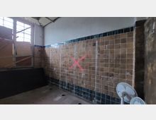 Ein leerer, überwiegend verlassener Raum mit braunen Fliesen an den Wänden, von denen einige beschädigt sind. Auf der Wand ist ein rotes "X" aufgesprüht, links befinden sich verbarrikadierte Fenster, und im Vordergrund ist ein weißer Standventilator sichtbar.