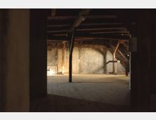 Ein leerer Dachboden mit Holzbalken und Holzboden, schwach beleuchtet durch einfallendes Tageslicht. Die Wände bestehen aus rohen Ziegeln oder verputztem Mauerwerk, und der Raum wirkt unrenoviert und rustikal.