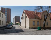 Straßenszene in einem Wohngebiet mit Kopfsteinpflaster, links und rechts von beigefarbenen Häusern mit roten Ziegeldächern gesäumt. Im Hintergrund stehen mehrere moderne mehrstöckige Gebäude, und parkende Autos sind entlang der Straße zu sehen.