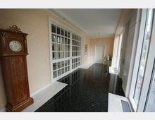 Heller Flur in einem eleganten Gebäude mit glänzendem schwarzen Steinboden, cremefarbenen Wänden und großen Fenstern auf der rechten Seite. Ein antikes Standuhrgehäuse steht links, und im Hintergrund befindet sich eine weiße Tür neben einer Glaswand mit zahlreichen Fenstereinsätzen.