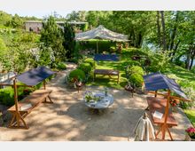 Grüne Gartenlandschaft mit schattigem Zeltbereich im Hintergrund, Holztischen mit blauen Sonnensegeln im Vordergrund und dekorativen Blumenbeeten sowie runden Büschen entlang der Wege. Im Zentrum steht ein runder Steintisch mit Blumenarrangements.