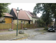 Straßenszene in einem Wohngebiet mit einem Einfamilienhaus im traditionellen Stil, roten Ziegeldächern und einem Holzzaun mit Backsteinpfosten. Im Vordergrund steht eine grüne Straßenlaterne, Pflastersteine, und ein geparkter schwarzer Kleinwagen am Straßenrand.