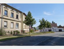 Straßenszene in einer Wohngegend mit mehrstöckigem Gebäude aus hellem Putz links, aufwändiger Fassadendekoration und mehreren Fenstern. Rechts stehen kleinere Wohnhäuser mit Satteldächern, davor sind Fußgängerwege, Bäume und ein Grünstreifen sichtbar; blauer Himmel im Hintergrund.