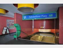 Eingangsbereich der U-Bahn-Station Fehrbelliner Platz (U3) in Berlin, mit roten Mosaikfliesen verkleideten Wänden und einem grünen Dekorelement. Ein blaues Stationsschild sowie Treppen, die in den Untergrund führen, sind sichtbar.