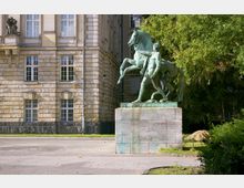 Eine Bronzestatue eines Mannes mit einem aufsteigenden Pferd, auf einem steinernen Sockel im Vordergrund, vor der Fassade eines historischen Gebäudes mit hellen Steinwänden und hohen Fenstern; im Hintergrund befinden sich Bäume.