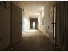 Verlassener Flur mit abblätternder Farbe an den Wänden, mehreren geöffneten Türen und einem leicht beschädigten Bodenbelag; am Ende des Flurs ist eine Glas-Notausgangstür mit Blick auf Tageslicht sichtbar.