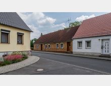 Straßenszene in einem Wohngebiet mit mehreren niedrigeren Häusern mit Satteldächern, darunter ein gelbes Haus mit Blumenbeet im Vordergrund, ein Haus mit verklinkerter Fassade und ein weiß verputztes Haus mit rotem Dach. Der Himmel ist leicht bewölkt, und ein Asphaltweg führt entlang der Gebäude.