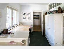 Helle Küche mit weißer Einrichtung, einschließlich eines großen Einbauschranks, eines Esstisches mit Eckbank und einem Fenster mit Sprossenglas. An der Wand hängen vier gerahmte Vogelzeichnungen, und im offenen Durchgang ist ein weiteres Gemälde sichtbar.