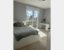 Helles Schlafzimmer mit grauen Wänden, einem Bett mit grauer Bettwäsche links und einem weißen Schreibtisch mit Dekoration in der Ecke rechts. Große Fenster mit Blick auf Häuser und Gärten lassen viel Tageslicht herein.