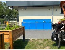 Ein Außenbereich mit erhöhten Holzbeeten im Vordergrund, einem gepflasterten Boden und einer Wand mit einer blauen Außenwaschstation. Rechts im Bild steht ein beladenes Fahrrad, im Hintergrund sind Bäume und ein Bauzaun zu sehen.