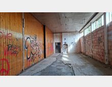 Verlassener, langer Innenraum einer Industriehalle mit hoher Betondecke, teilweise zerstörtem Boden und Graffiti an einer Holzvertäfelung sowie an Backsteinwänden. Rechts fällt Tageslicht durch eine Reihe hoher Fenster; am Ende führt eine Türöffnung zu einer Treppe.