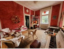 Wohnraum mit roten Wänden und Holzboden, eingerichtet mit einem runden weißen Tisch und geflochtenen Stühlen, dekoriert mit schaffellartigen Überzügen. Im Hintergrund ein großer Schreibtisch vor einem Fenster mit Blick ins Grüne, rechts ein weißes Klavier und diverse Regalflächen.