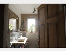 Holzbad mit natürlichem Licht, das durch ein großes Fenster mit Blick auf Pflanzen fällt. Der Raum enthält ein Waschbecken auf einer Holzoberfläche, Fliesen an der Wand, eine Wandspiegel und Dekorationen wie Zimmerpflanzen.