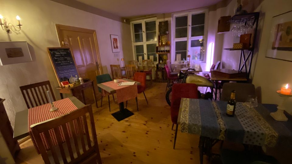 Gemütlicher Gastraum eines kleinen Cafés mit Holzdielenboden, mehreren kleinen Tischen mit gemusterten Tischdecken und gemischten Stühlen. Hinten stehen hohe Fenster, ein Schrankregal und eine Kreidetafel; an der Wand sorgen Kerzen und eine indirekte Lichtquelle für warmes Licht.