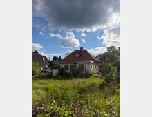 Einfamilienhaus mit rotem Ziegeldach und Schornstein in einem verwilderten Garten mit hohem Gras und dichtem Bewuchs. Im Hintergrund stehen weitere Wohnhäuser und Bäume unter einem teils bewölkten Himmel.