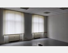 Leerer Innenraum mit zwei großen Fenstern, die durch vertikale weiße Jalousien bedeckt sind. Die Wände sind weiß gestrichen, die Decke ist mit Rasterplatten versehen, und unter den Fenstern befinden sich zwei Heizkörper.