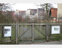Holztor mit Lattenzaun, das den Eingang zum Comenius-Garten markiert, mit einem Briefkasten und einer Infotafel. Im Hintergrund sind grasbewachsene Flächen, kahle Bäume und Wohnhäuser mit roten Ziegeldächern zu sehen.