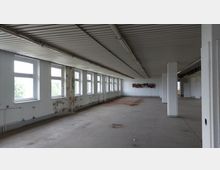 Ein leerer, industriell wirkender Innenraum mit einer niedrigen Decke aus Metallplatten und mehreren Reihen rechteckiger Fenster an der linken Wand. Der Boden ist aus Beton, teilweise verschmutzt, und die Wände zeigen Abnutzungsspuren; weiße Säulen teilen den Raum.