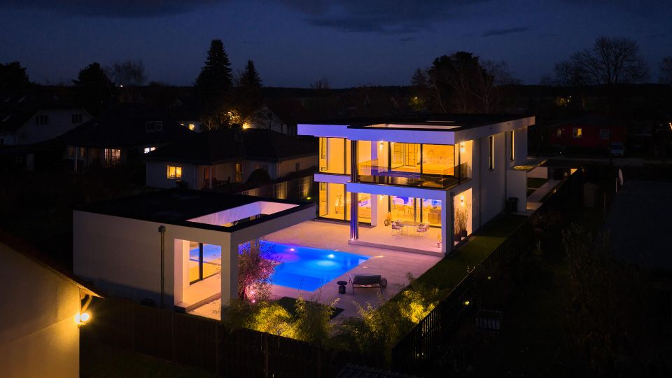 Nachtaufnahme eines modernen, zweigeschossigen Flachdachhauses mit großen Glasfronten und Balkon, angrenzend an eine beleuchtete Terrasse mit rechteckigem Swimmingpool. Das Anwesen ist eingezäunt und liegt in einem Wohngebiet mit umliegenden Häusern und Bäumen im Hintergrund.