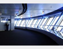 Innenansicht einer modernen Aussichtsplattform mit gebogenen, bodentiefen Fenstern, die von blauer Beleuchtung Akzente erhalten. Ein Fernglas und eine große Vase mit Blumen sind neben metallischen Geländern sichtbar.