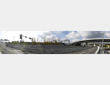 Eine mehrspurige Autobahnauffahrt mit geteerten Fahrbahnen und Betonschutzwänden, flankiert von kahlen Bäumen und Verkehrsschildern. Im Hintergrund ist eine geschwungene Überführung sichtbar, ergänzt durch gelbe Herbstbäume und einen bewölkten Himmel.