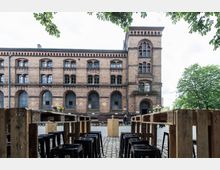 Backstein-Gebäude im historischen Stil mit mehreren Stockwerken, großen Bogenfenstern und verzierten Fassadenelementen. Im Vordergrund stehen Tische und Hocker aus Holzpaletten auf einem gepflasterten Hof, gesäumt von Bäumen.