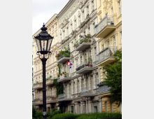 Rundbogenfenster, Balkone mit Pflanzen sowie verziertes Stuckdekor prägen das historische Mehrfamilienhaus in einer Stadtstraße. Im Vordergrund steht eine schwarze, nostalgische Straßenlaterne, umgeben von Grünpflanzen.