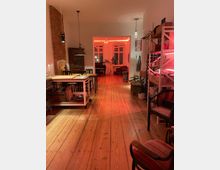 Ein offener Wohnraum mit Holzdielenboden, einer Sichtziegelwand links und einer Kücheninsel in der Mitte. Im Hintergrund ein angrenzendes Zimmer mit roten Akzentlichtern, großen Fenstern und Pflanzen; Regale und Stühle stehen rechts im Vordergrund.