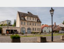 Straßenszene mit einem historischen Gebäude mit der Aufschrift "Rechtsanwälte" im Zentrum. Daneben befinden sich ein Textilgeschäft sowie kleinere Wohnhäuser mit roten und grauen Dächern, umgeben von einer mit Pflastersteinen bedeckten Straße, Laternen und Pflanzkübeln.