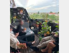 Ein Balkon mit Holzboden, umgeben von zahlreichen Topfpflanzen und Blumen, teils an Regalen und Geländern arrangiert. Im Hintergrund ist der Ausblick auf eine städtische Umgebung mit Bäumen und Häusern zu sehen; im Vordergrund sitzen mehrere Personen auf einer Sitzgruppe mit Kissen.